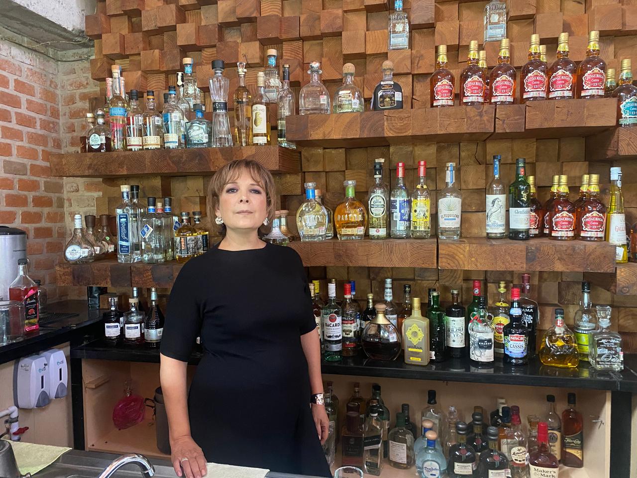 Eine Frau mit braunen Haaren und schwarzem Kleid steht an einer Bar. Hinter ihr ein Holzregal voll mit bunten Flaschen: die Sommeliére Ana Maria Romero.