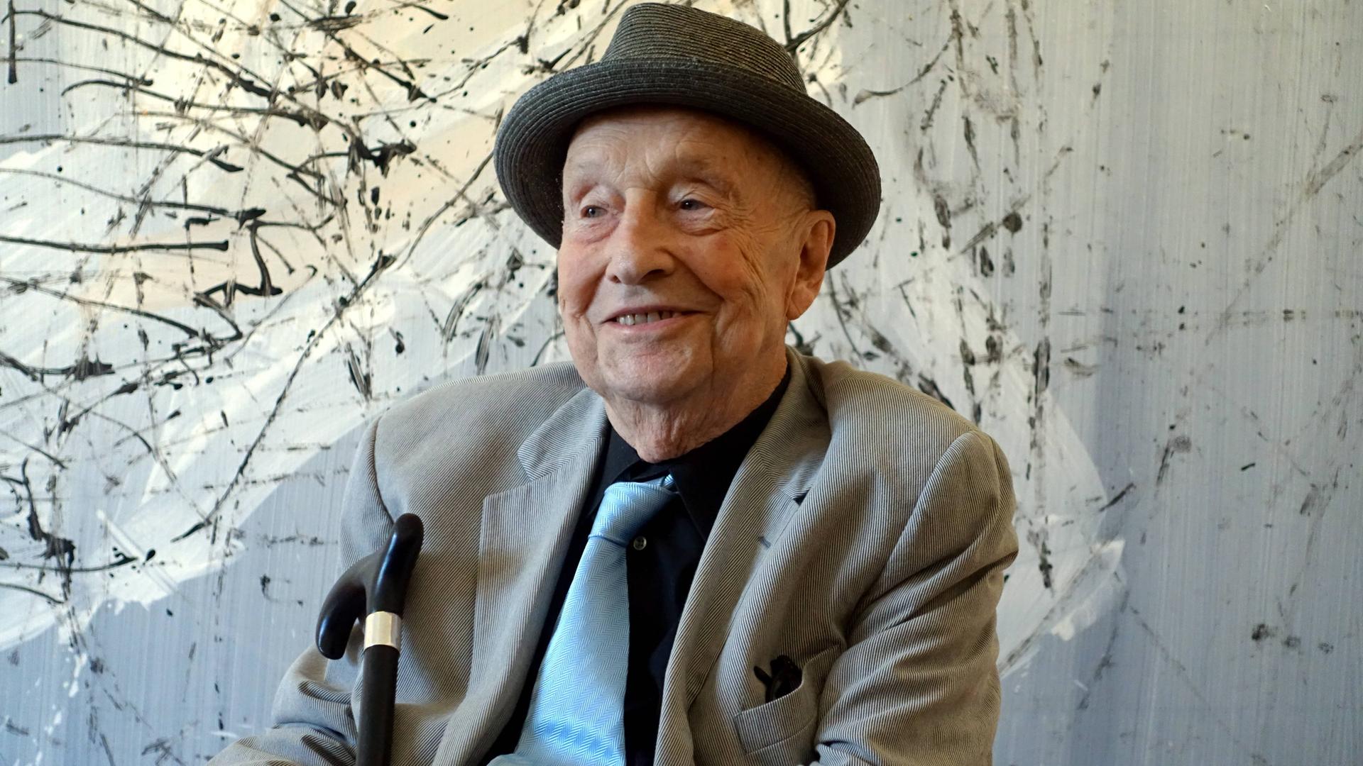 Georg Baselitz mit Hut vor einer abstrakten Malerei. Der Künstler lächelt