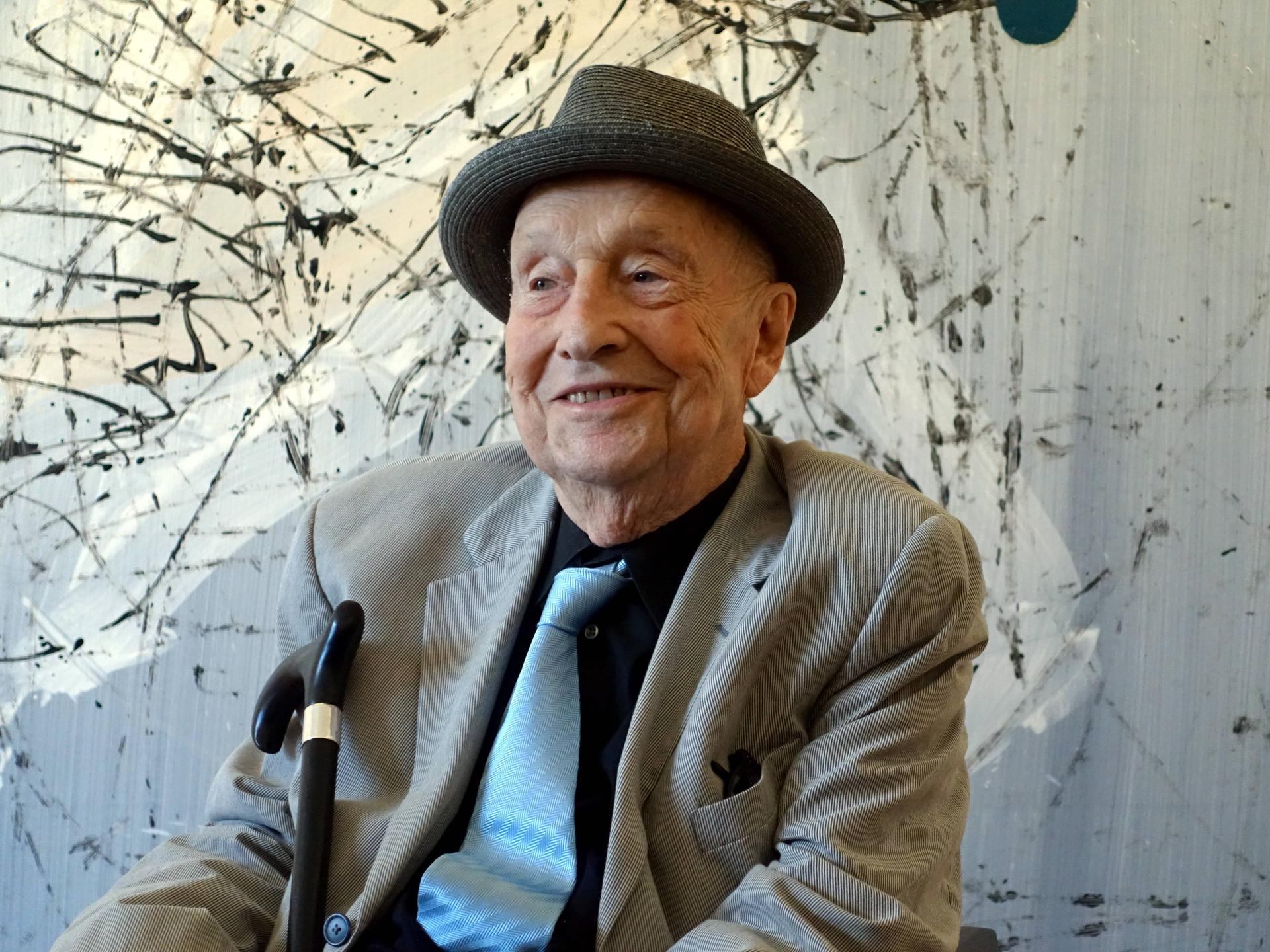 Georg Baselitz mit Hut vor einer abstrakten Malerei. Der Künstler lächelt