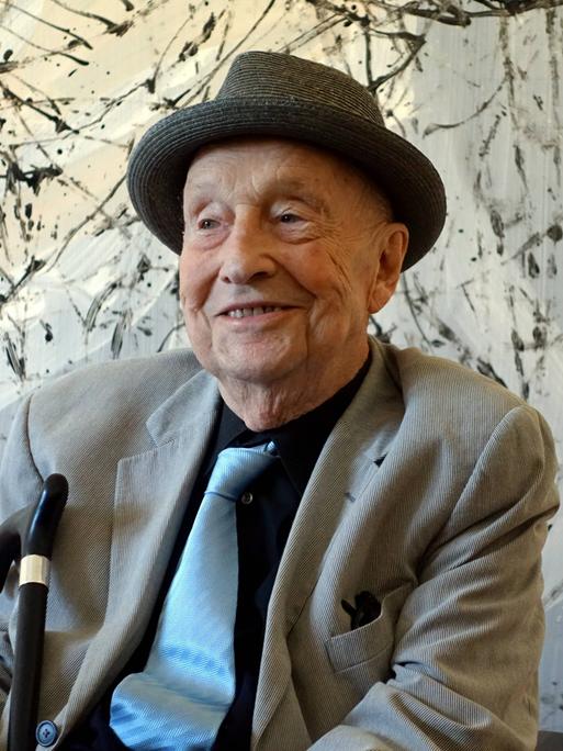 Georg Baselitz mit Hut vor einer abstrakten Malerei. Der Künstler lächelt
