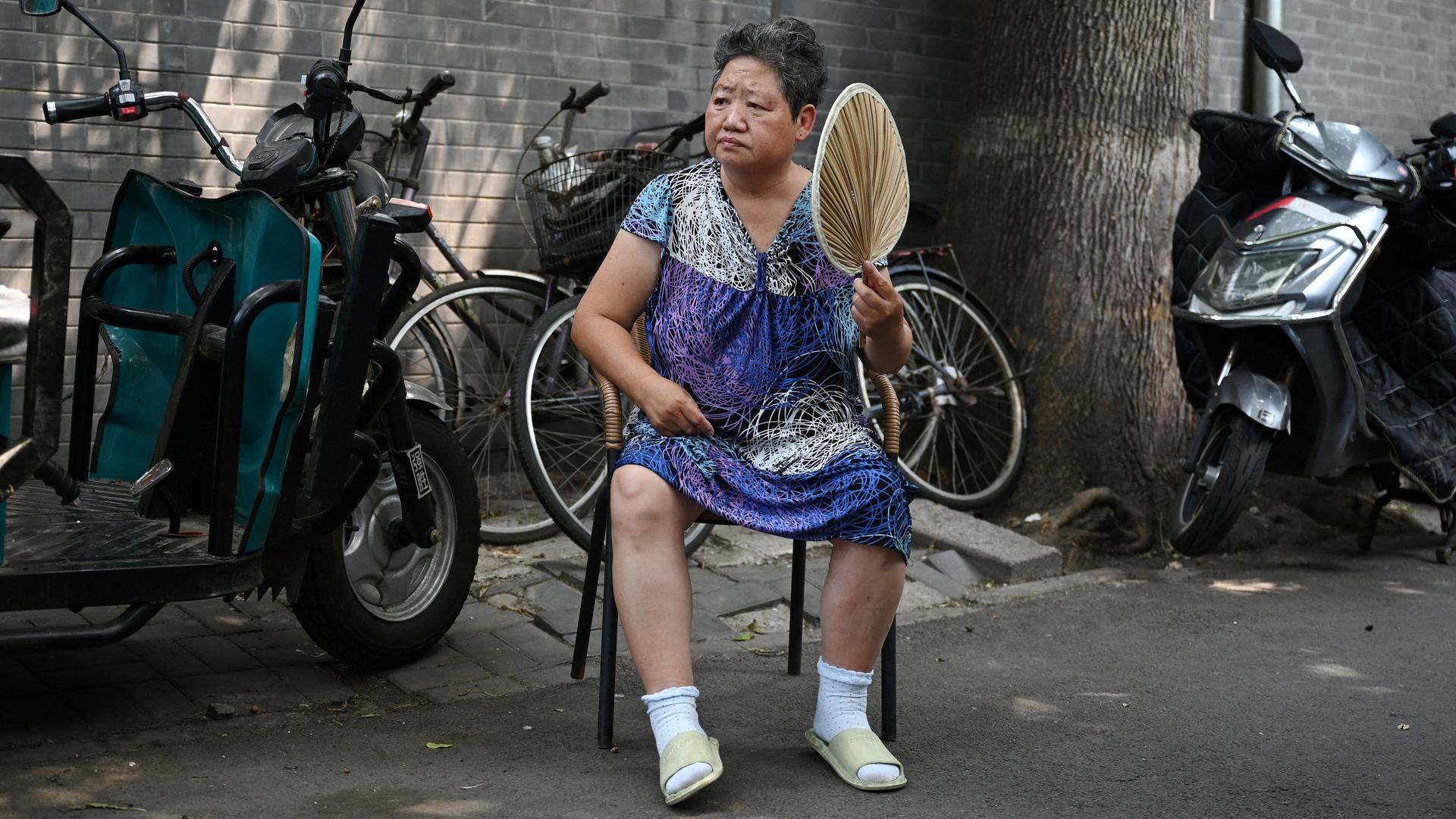 Eine chinesische Frau sitzt auf einem Stuhl, sie hält einen Fächer in der Hand und schaut erschöpft.