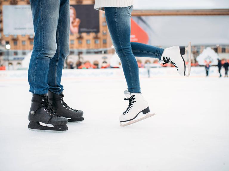 Man sieht die Beine von einem Mann und einer Frau, die auf Schlittschuhen auf dem Eis stehen. 