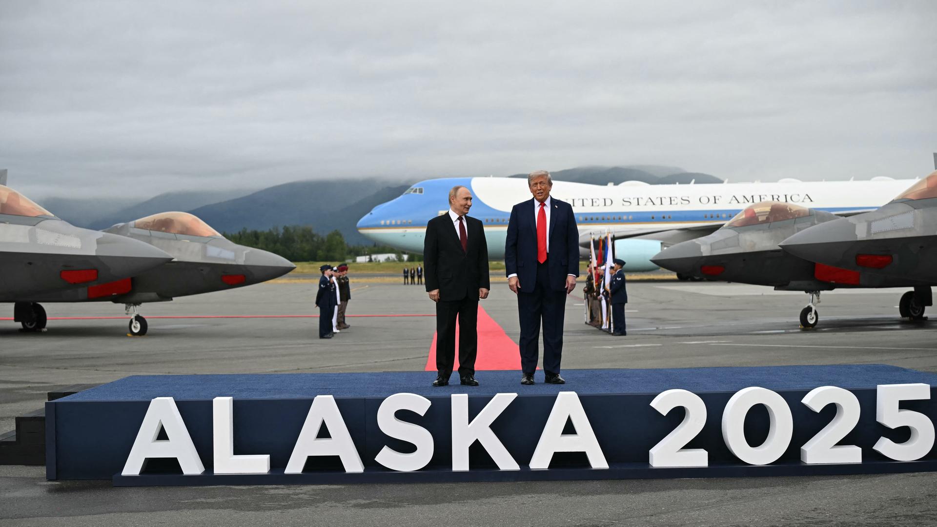Trump und Putin stehen auf einem Podest, auf dem der Schriftzug "Alaska 2025" angebracht ist. Im Hintergrund stehen Flugzeuge.