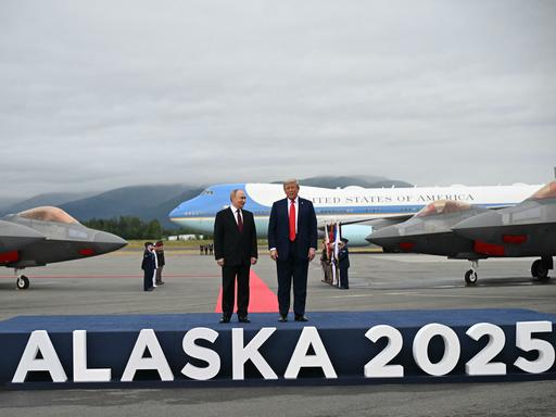 Trump und Putin stehen auf einem Podest, auf dem der Schriftzug "Alaska 2025" angebracht ist. Im Hintergrund stehen Flugzeuge.