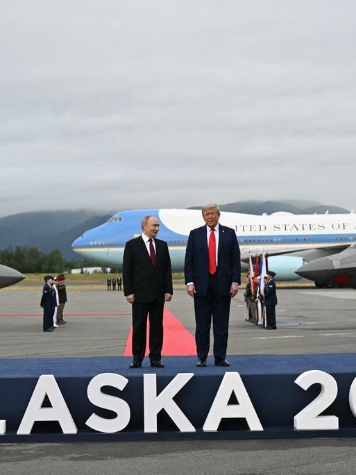Trump und Putin stehen auf einem Podest, auf dem der Schriftzug "Alaska 2025" angebracht ist. Im Hintergrund stehen Flugzeuge.