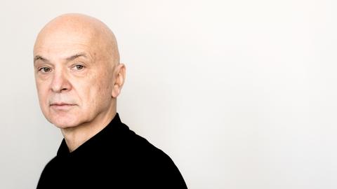 Ein Mann steht asymmetrisch im Bild vor weiße Hintergrund und schaut in die Kamera