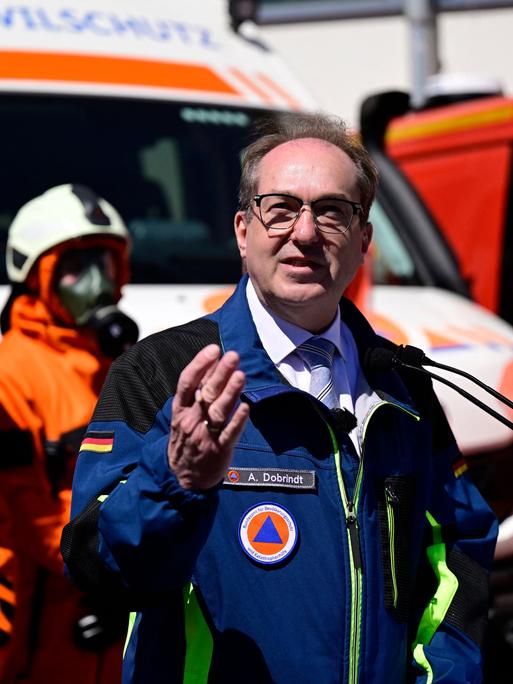Alexander Dobrindt steht an einem Rednerpult und spricht. Im Hintergrund sind Personen in Schutzkleidung vom THW zu sehen.
