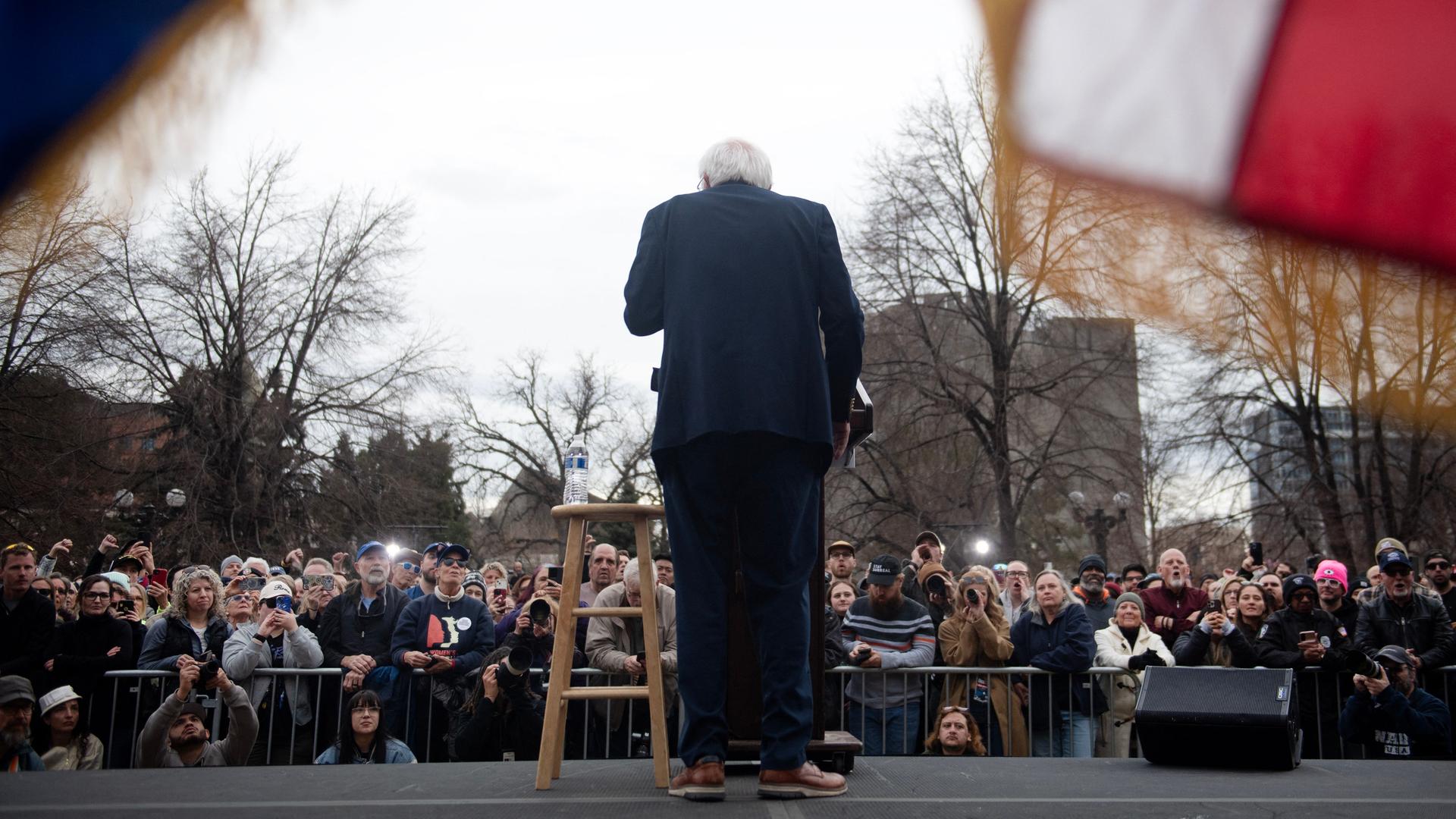 Rückansicht von Bernie Sanders, der während einer Ansprache vor Publikum an einem Pult steht. Rückansicht von Bernie Sanders, der während einer Ansprache vor Publikum an einem Pult steht.