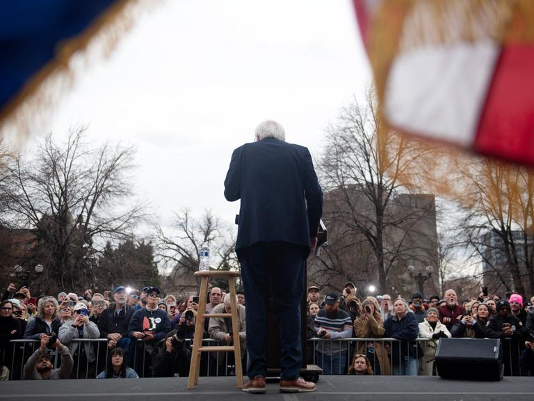 Rückansicht von Bernie Sanders, der während einer Ansprache vor Publikum an einem Pult steht.