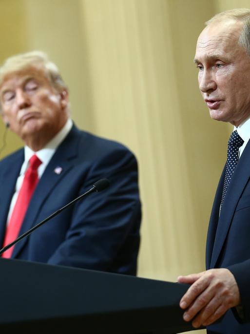 US-Präsident Trump und Russlands Präsident Putin geben gemeinsam eine Pressekonferenz: Putin steht am Redepult und spricht, Trump dreht sich zu ihm und hört zu.