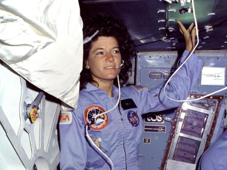 Sally Ride trägt einen blauen Overal, während sie sich auf einer Raumstation mit Headset aufhält.