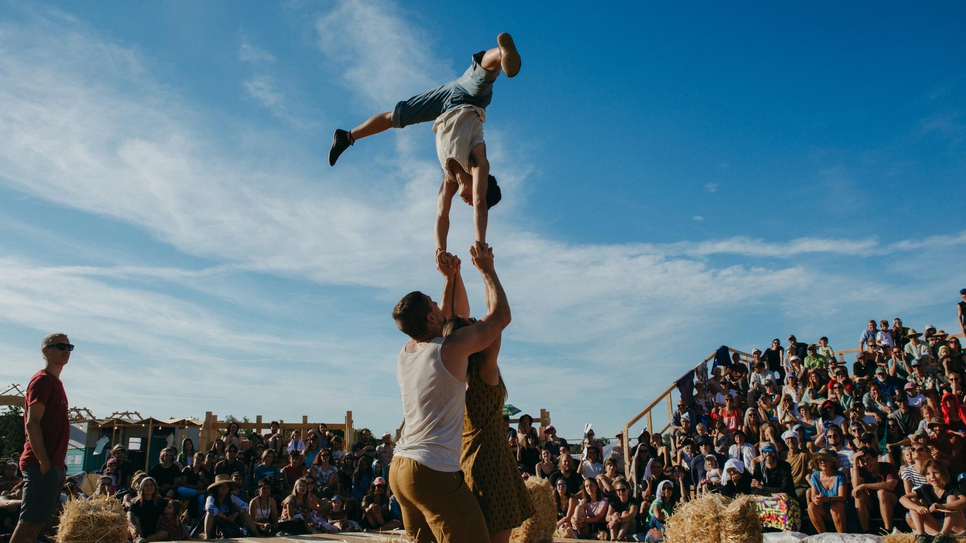 Auf einer Open-Air-Bühne macht ein Mann einen Handstand auf den Händen einer Artistin und eines Artisten. Auf einer Open-Air-Bühne macht ein Mann einen Handstand auf den Händen einer Artistin und eines Artisten.