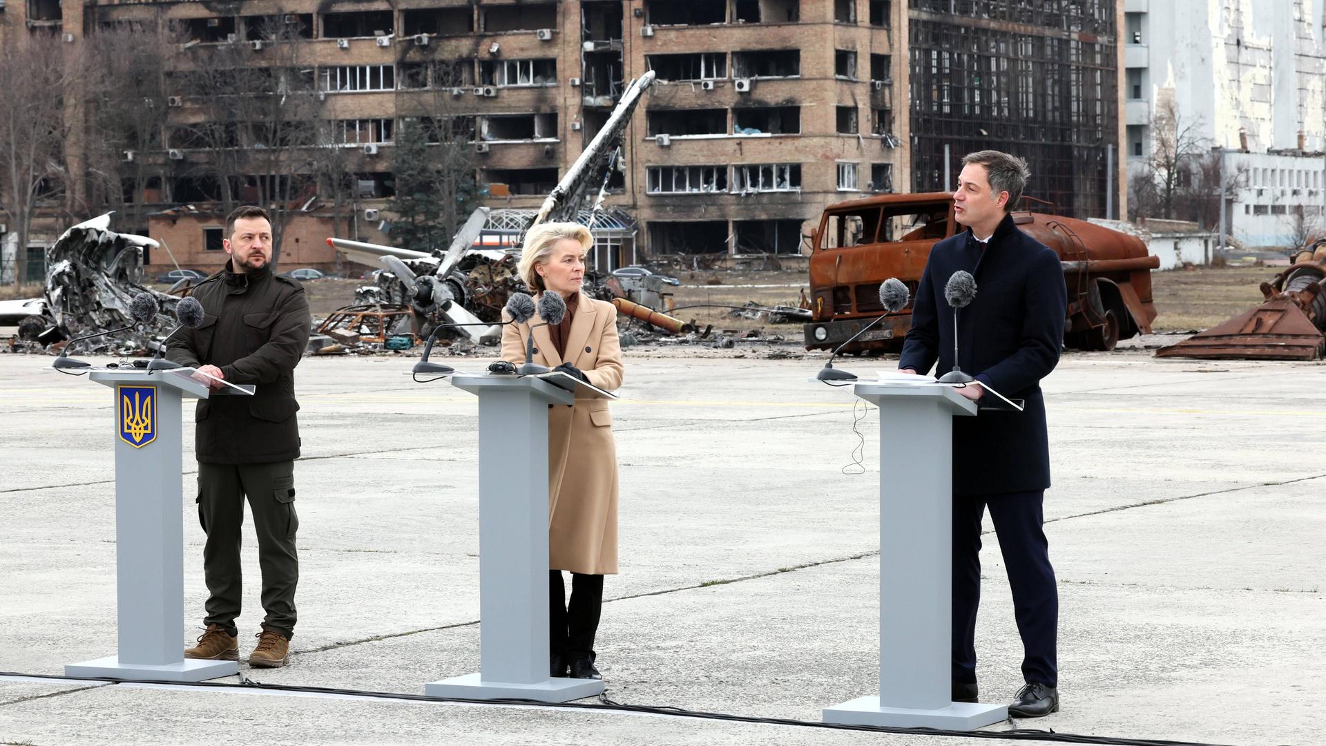 Der ukrainische Präsident Selenskyj, EU-Kommissionspräsidentin Von der Leyen und der belgische Ministerpräsident De Croo stehen in Kiew nebeneinander an Rednerpulten. Im Hintergrund ist ein zerstörtes Gebäude zu sehen.