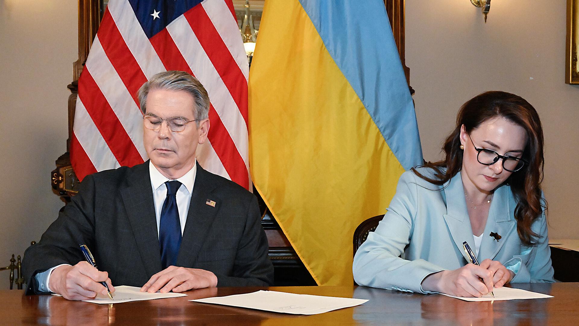 US-Finanzminister Bessent und die ukrainische Wirtschaftsministerin Swyrydenko unterschreiben das Rohstoffabkommen. Bessent (li) hat kurze graue Haare und sitzt vor der US-Flagge. Swyrydenko (re) vor einer ukrainischen Flagge.