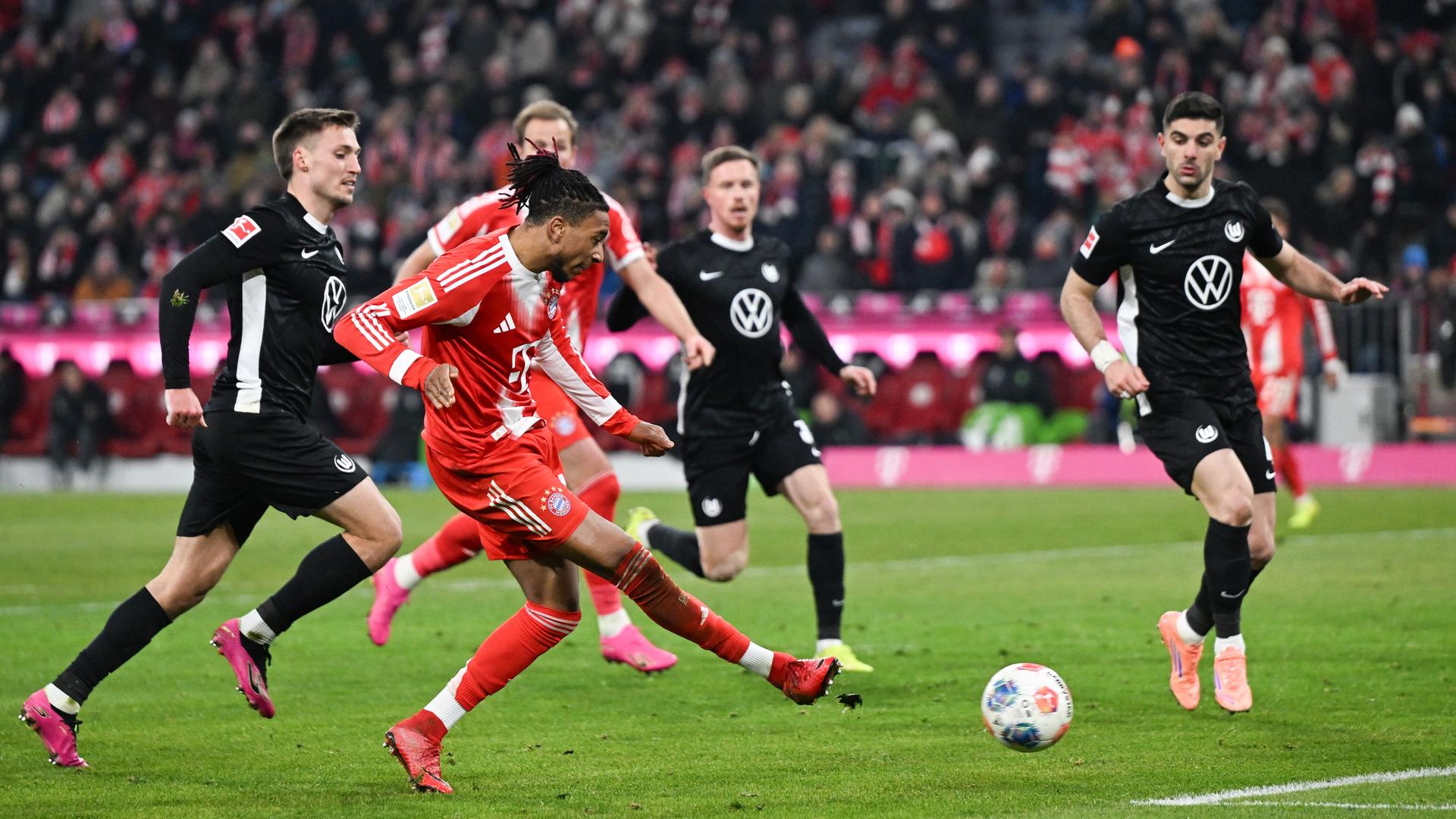 Michael Olise im roten Bayern-Trikot ist umgeben von Wolfsburger Spielern im schwarzen Trikot. Olise spielt den Ball zum 7:1 für die Bayern. 
