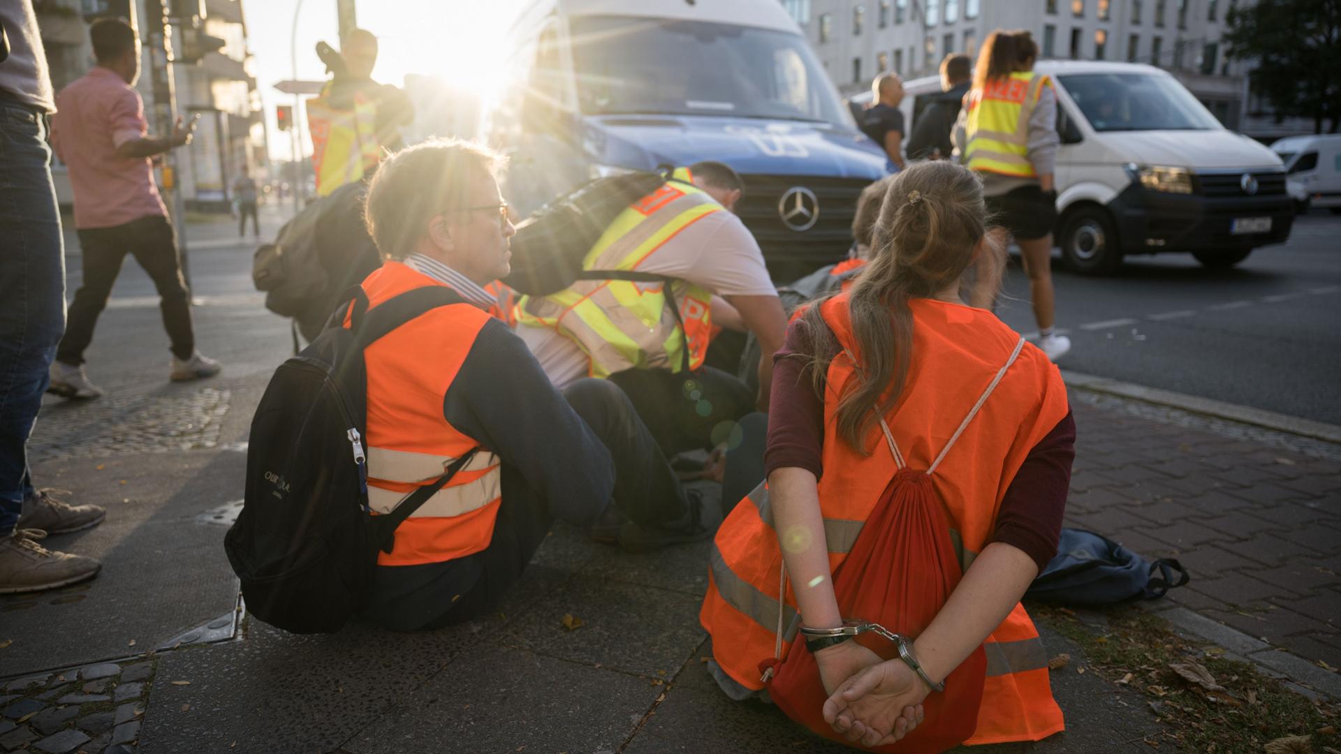 Berlin: Aktivisten der Klimaschutzgruppe "Letzte Generation" sitzen nach einer Blockade am Spandauer Damm in Handschellen an einem Polizeiwagen. Berlin: Aktivisten der Klimaschutzgruppe "Letzte Generation" sitzen nach einer Blockade am Spandauer Damm in Handschellen an einem Polizeiwagen.