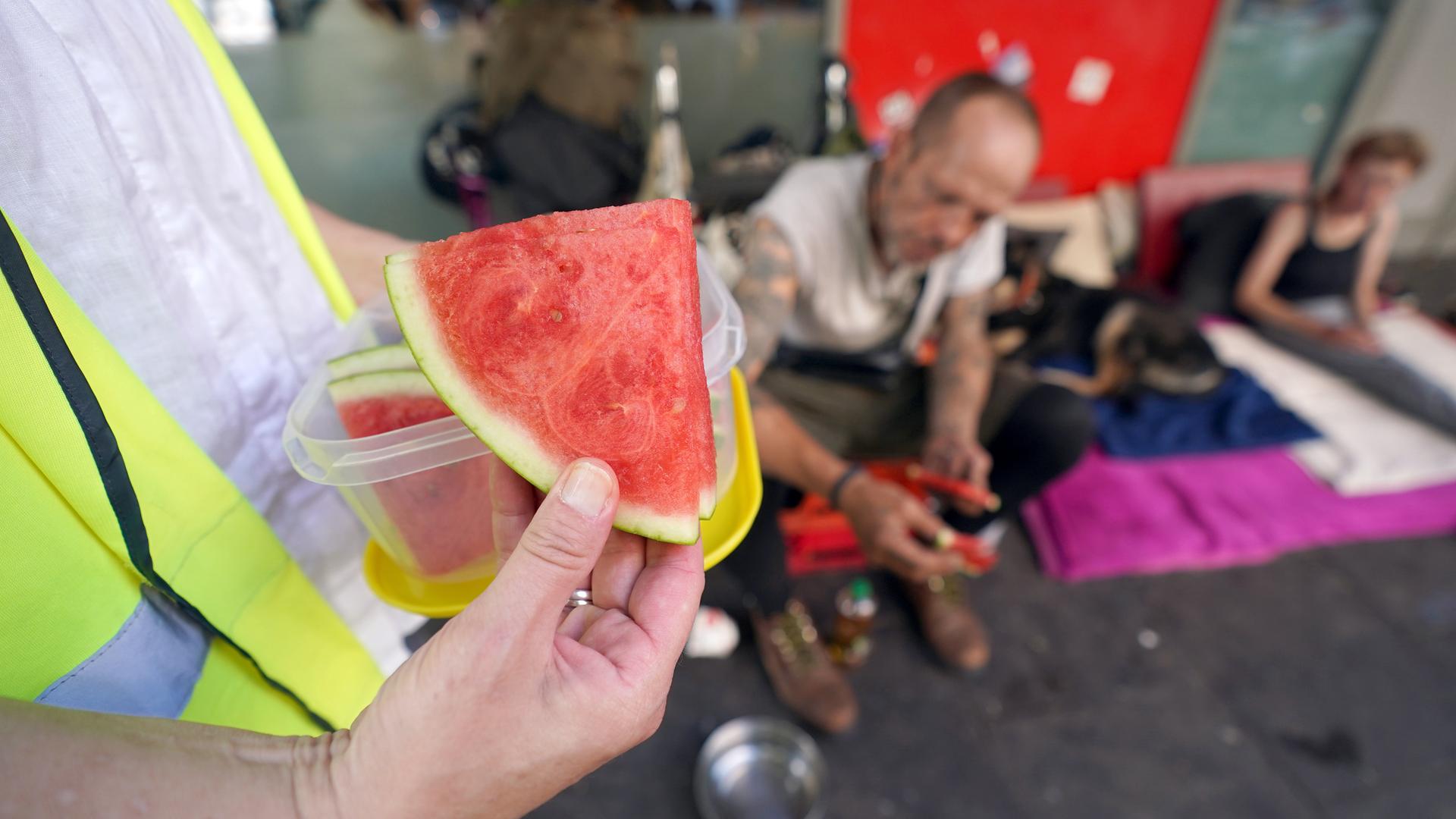 Nahaufnahme einer Hand, die Wassermelonenstücke an Obdachlose austeilt, die im Hintergrund unscharf zu sehen sind.