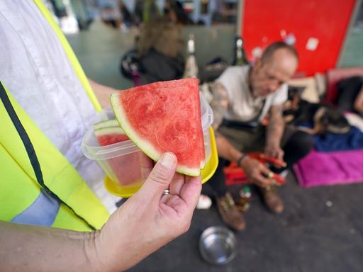 Nahaufnahme einer Hand, die Wassermelonenstücke an Obdachlose austeilt, die im Hintergrund unscharf zu sehen sind.