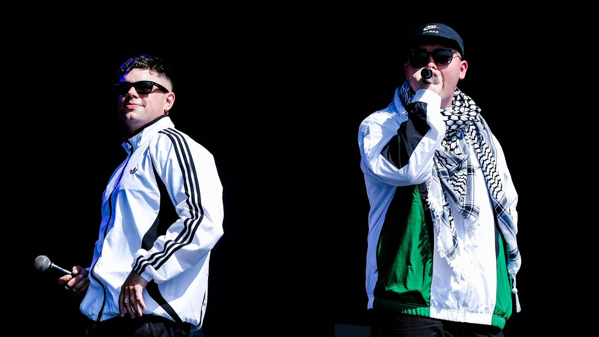 Zwei weiße Rapper in Trainingsanzügen und Sonnenbrillen auf einer Bühne. Der Mann rechts trägt eine Käppi und einen Palästinenserschal. Zwei weiße Rapper in Trainingsanzügen und Sonnenbrillen auf einer Bühne. Der Mann rechts trägt eine Käppi und einen Palästinenserschal.