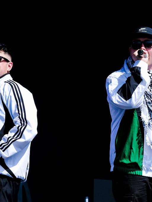 Zwei weiße Rapper in Trainingsanzügen und Sonnenbrillen auf einer Bühne. Der Mann rechts trägt eine Käppi und einen Palästinenserschal. (Quelle: imago)