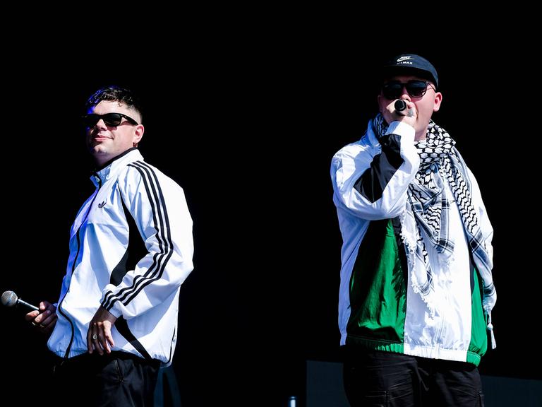 Zwei weiße Rapper in Trainingsanzügen und Sonnenbrillen auf einer Bühne. Der Mann rechts trägt eine Käppi und einen Palästinenserschal. (Quelle: imago)