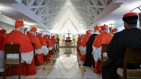 Bischöfe sitzen auf Stühlen in einer Halle mit spitzen Dach. An deren Ende steht ein Altar mit Jesus-Kreuz. 
