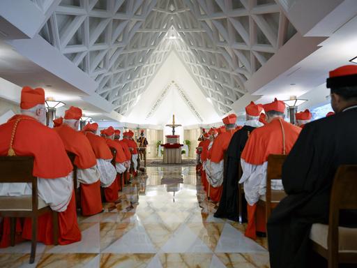 Bischöfe sitzen auf Stühlen in einer Halle mit spitzen Dach. An deren Ende steht ein Altar mit Jesus-Kreuz. 