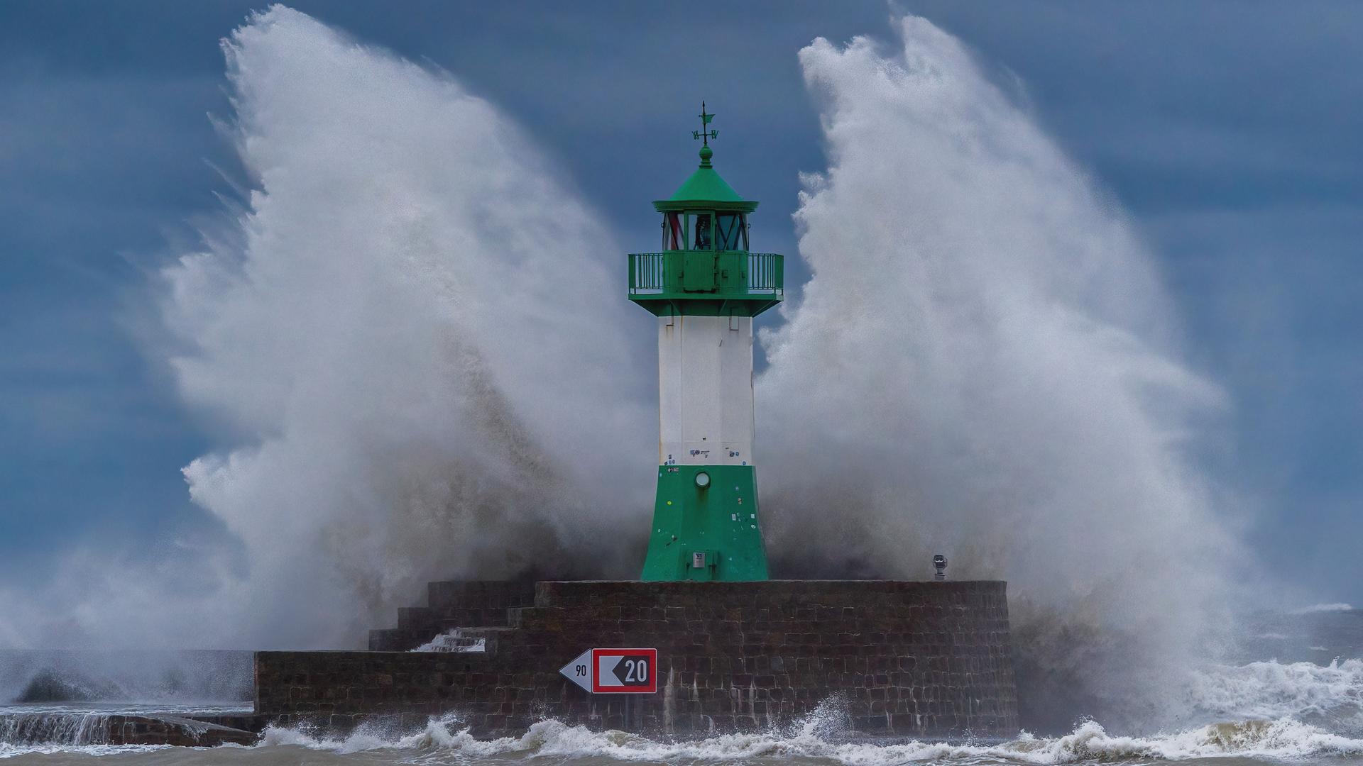 Hohe Wellen schlagen gegen einen grün-weißen Leuchtturm auf der Ostmole an der Ostseeküste, wo sich im Oktober 2023 eine schwere Sturmflut ereignete.