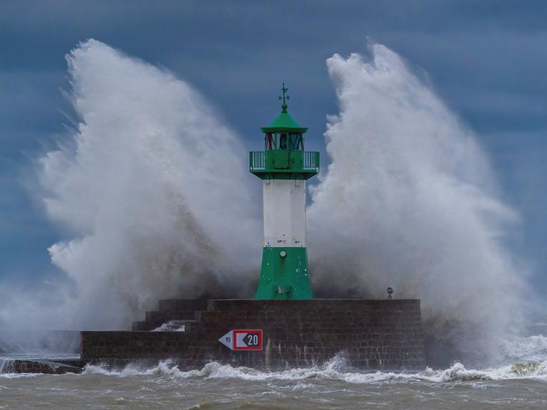 Hohe Wellen schlagen gegen einen grün-weißen Leuchtturm auf der Ostmole an der Ostseeküste, wo sich im Oktober 2023 eine schwere Sturmflut ereignete.