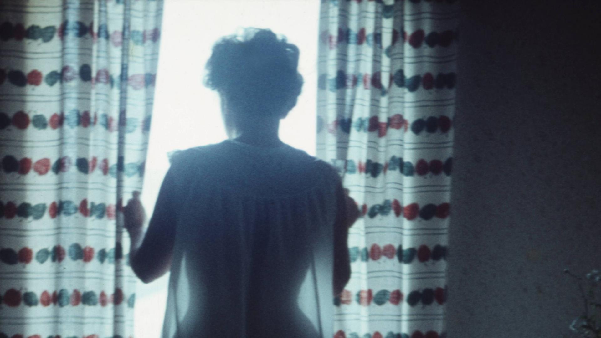 Aufnahme von Mitte der 1950er-Jahre. Eine Frau steht an einem Fenster durch das gleissendes Licht fällt. Von ihr sind nur schemenhaft Umrisse zu erkennen. Sie zieht die Gardinen auseinander. Rechts neben ihr steht ein Küchentisch.