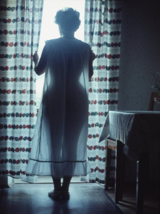 Aufnahme von Mitte der 1950er-Jahre. Eine Frau steht an einem Fenster durch das gleissendes Licht fällt. Von ihr sind nur schemenhaft Umrisse zu erkennen. Sie zieht die Gardinen auseinander. Rechts neben ihr steht ein Küchentisch.
