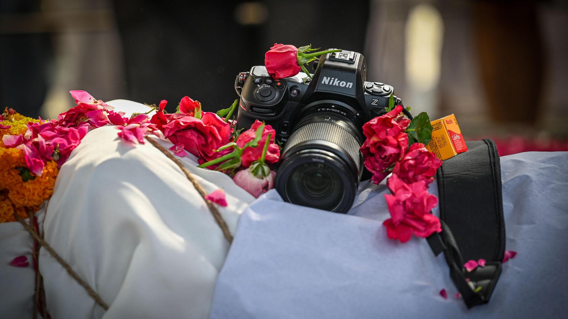 Eine Nikon-Kamera liegt auf dem aufgebahrten Körper des Toten, von dem nur ein weißes Tuch zu sehen ist und Blumen rund um die Kamera