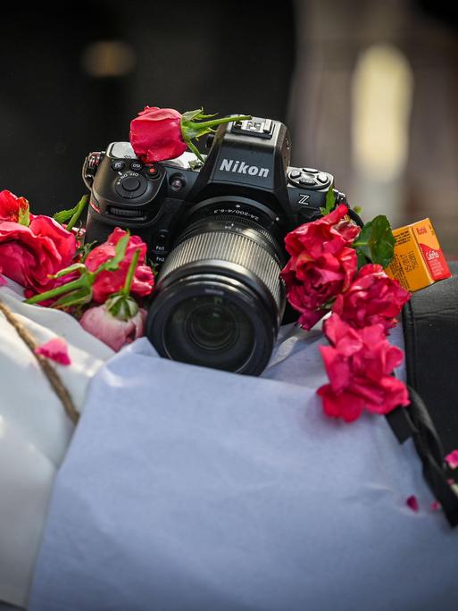 Eine Nikon-Kamera liegt auf dem aufgebahrten Körper des Toten, von dem nur ein weißes Tuch zu sehen ist und Blumen rund um die Kamera