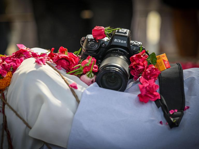 Eine Nikon-Kamera liegt auf dem aufgebahrten Körper des Toten, von dem nur ein weißes Tuch zu sehen ist und Blumen rund um die Kamera