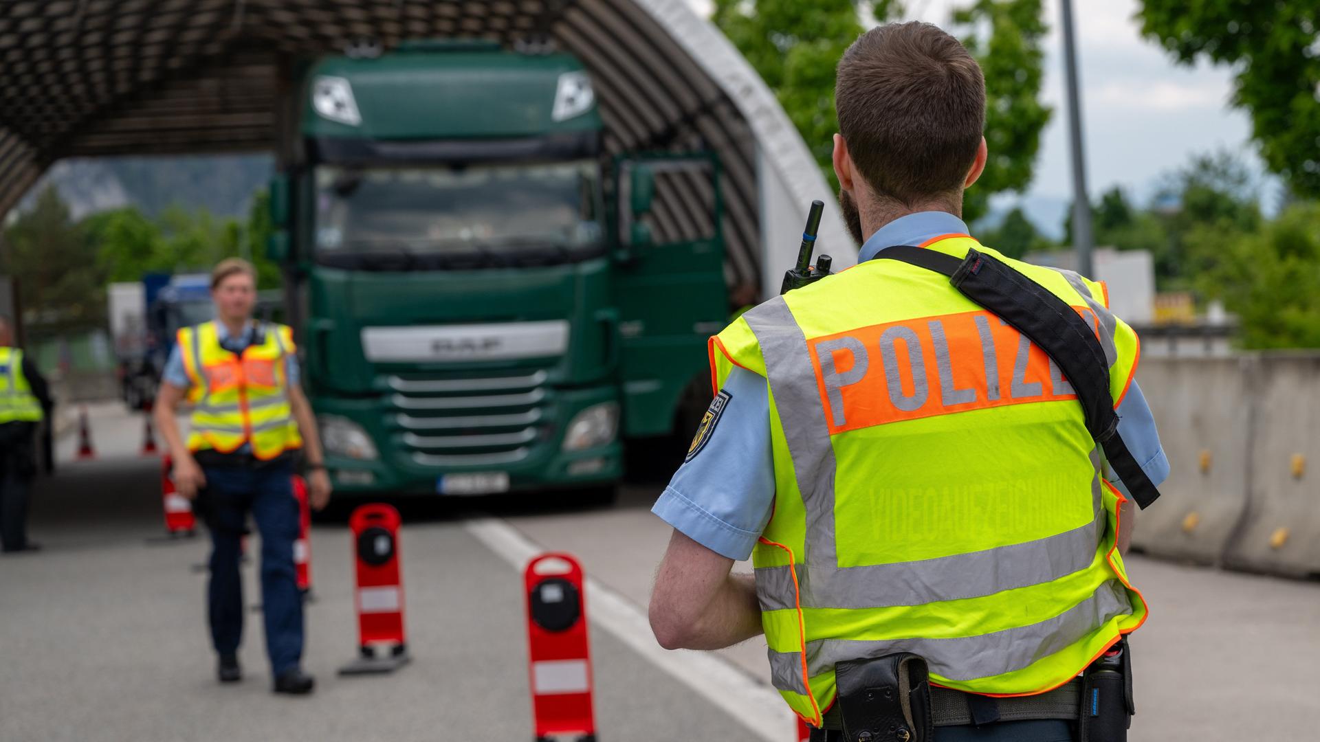 Zwei Polizisten in gelben Warnwesten stehen an einer mit Signalposten verengten Straße. Im Hintergrund zwei LKW.