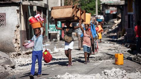 Szene aus der haitianischen Hauptstadt Port-au-Prince: Mehrere Personen gehen mit Gegenständen auf dem Kopf eine zerstörte Straße voller Schutt entlang.