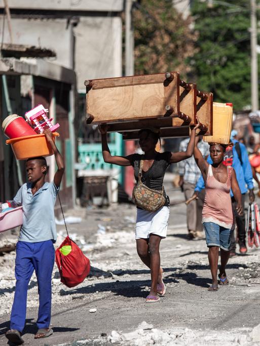 Szene aus der haitianischen Hauptstadt Port-au-Prince: Mehrere Personen gehen mit Gegenständen auf dem Kopf eine zerstörte Straße voller Schutt entlang.