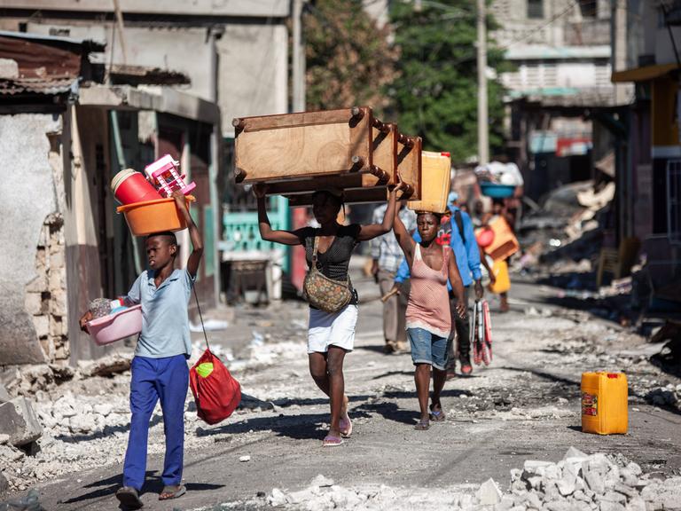 Szene aus der haitianischen Hauptstadt Port-au-Prince: Mehrere Personen gehen mit Gegenständen auf dem Kopf eine zerstörte Straße voller Schutt entlang.