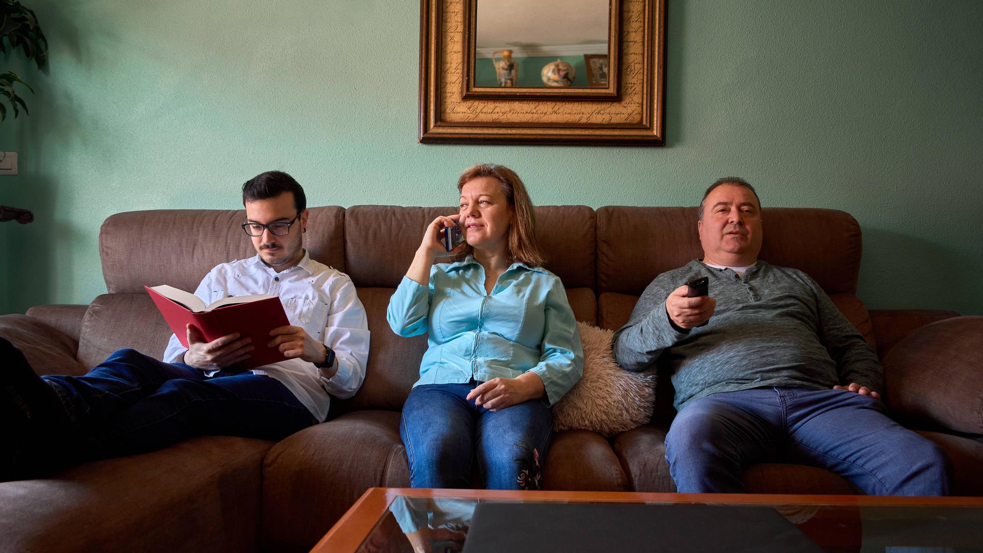 Eine Familie sitzt auf der Couch. Der erwachsene Sohn liest in einem Buch, die Mutter telefoniert, der Vater bedient eine Fernbedienung und sieht offenbar in den Fernseher. 