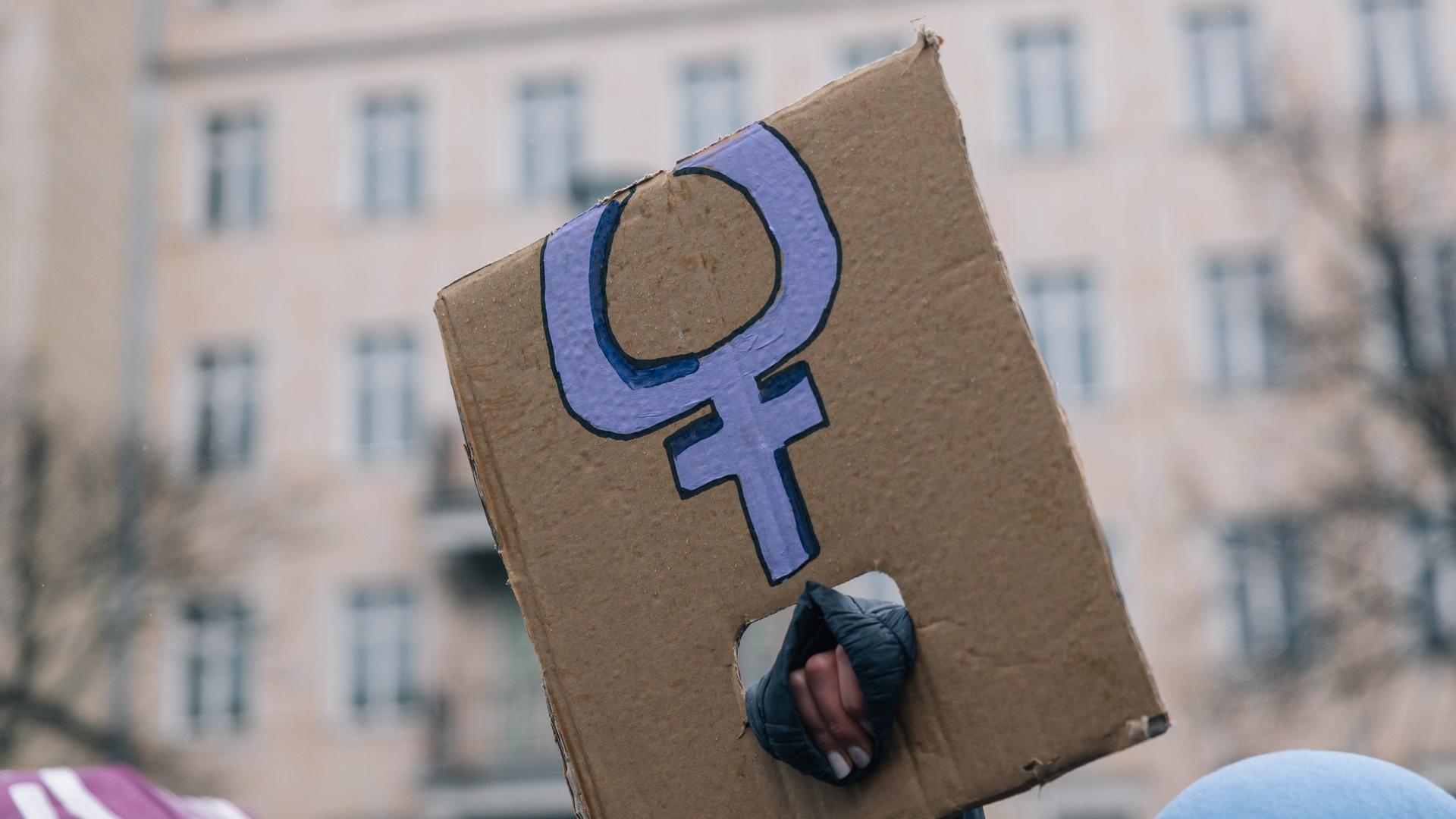 Eine Hand, die ein abgeknicktes Transparent mit dem violetten Symbol für Frauen hält Eine Hand, die ein abgeknicktes Transparent mit dem violetten Symbol für Frauen hält