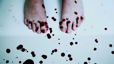 Blick auf zwei nackte Füße auf weißem Boden, darauf und darum herum Bluttropfen. Blick auf zwei nackte Füße auf weißem Boden, darauf und darum herum Bluttropfen.