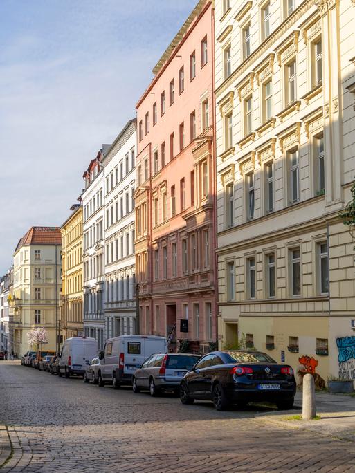 Eine Straße mit schönen Altbauwohnungen in Berlin. Auf der Straße parken ein paar Autos. Vor einem Haus im Vordergrund ist auch ein kleiner, grüner Baum oder Strauch.