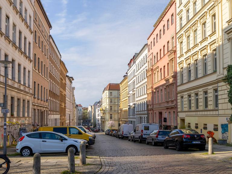 Eine Straße mit schönen Altbauwohnungen in Berlin. Auf der Straße parken ein paar Autos. Vor einem Haus im Vordergrund ist auch ein kleiner, grüner Baum oder Strauch.