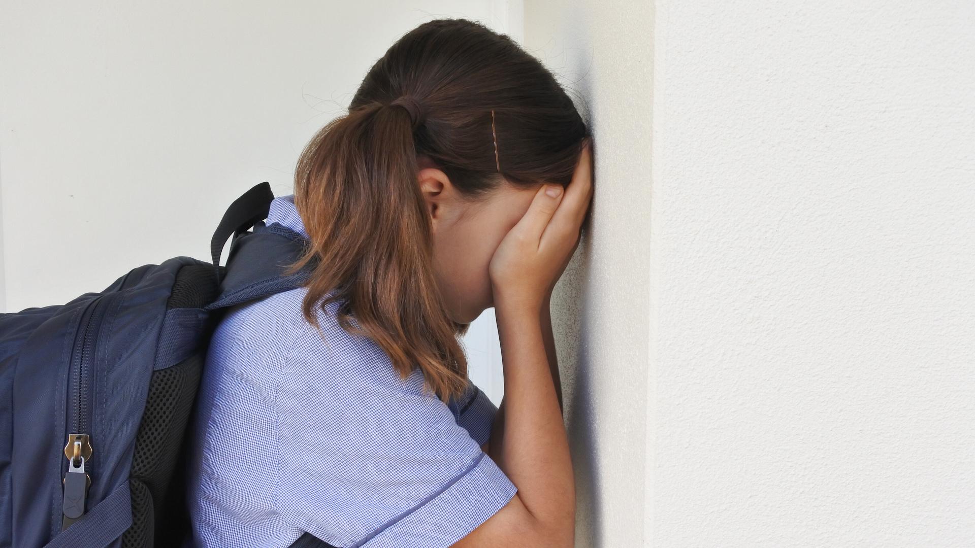 Ein junges Mädchen steht an einer Wand. Es hält die Hände vor sein Gesicht.