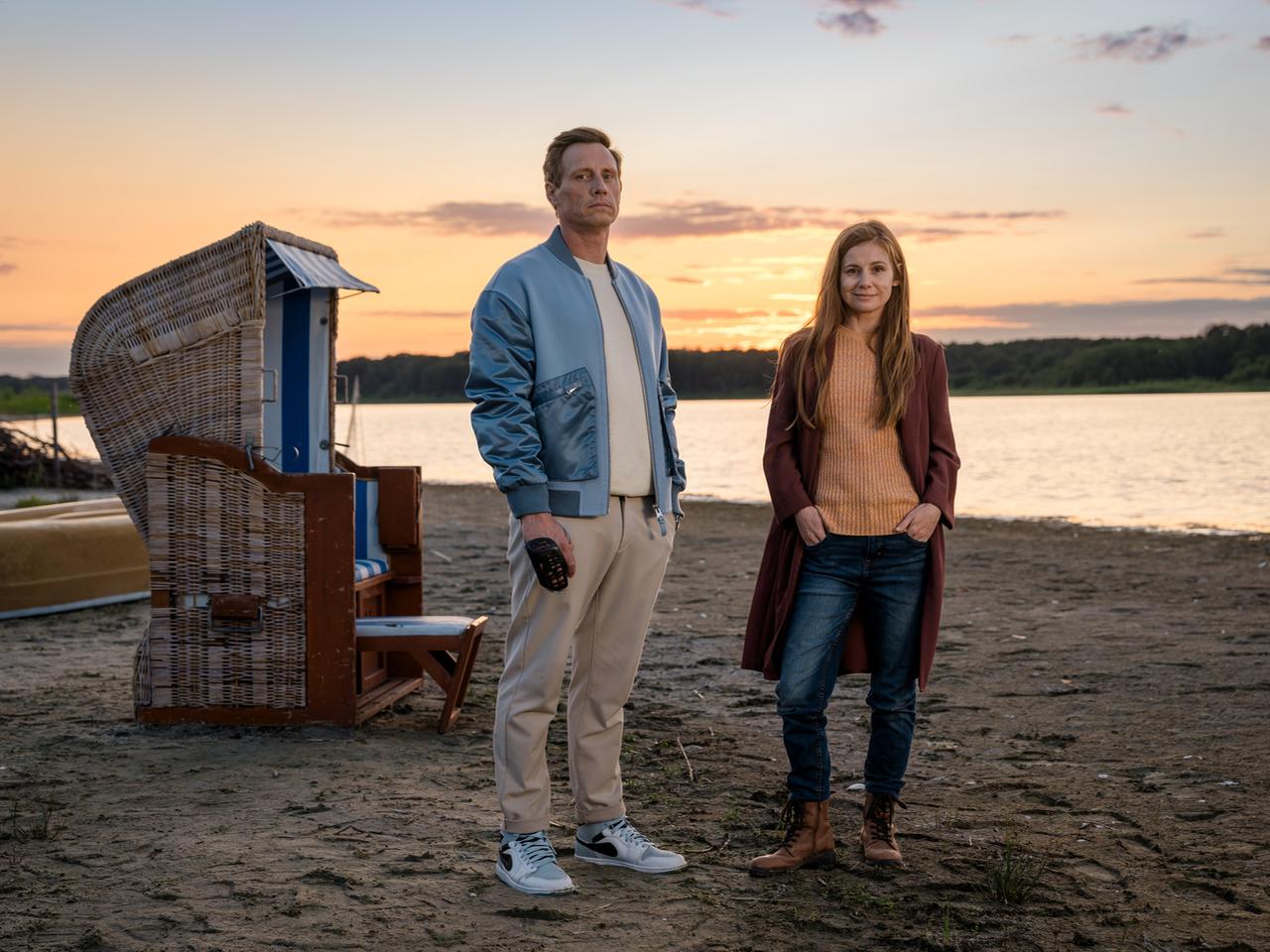 Ein Mann und eine Frau stehen in der Abenddämmerung an einem Strand. Neben ihnen ein Strandkorb, hinter dem Wasser liegt Wald. Er hat eine blaue Jacke über einem weißen Shirt, sie trägt eine rote Jacke über einem orangefarbenen Pullover. 