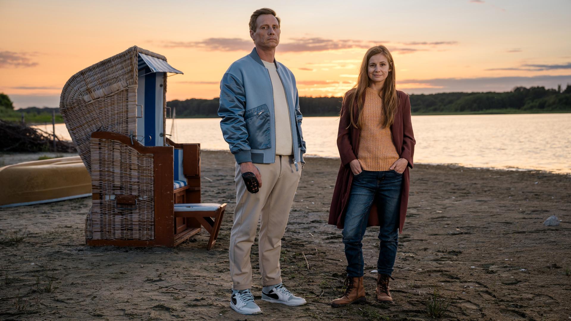 Ein Mann und eine Frau stehen in der Abenddämmerung an einem Strand. Neben ihnen ein Strandkorb, hinter dem Wasser liegt Wald. Er hat eine blaue Jacke über einem weißen Shirt, sie trägt eine rote Jacke über einem orangefarbenen Pullover. Ein Mann und eine Frau stehen in der Abenddämmerung an einem Strand. Neben ihnen ein Strandkorb, hinter dem Wasser liegt Wald. Er hat eine blaue Jacke über einem weißen Shirt, sie trägt eine rote Jacke über einem orangefarbenen Pullover.