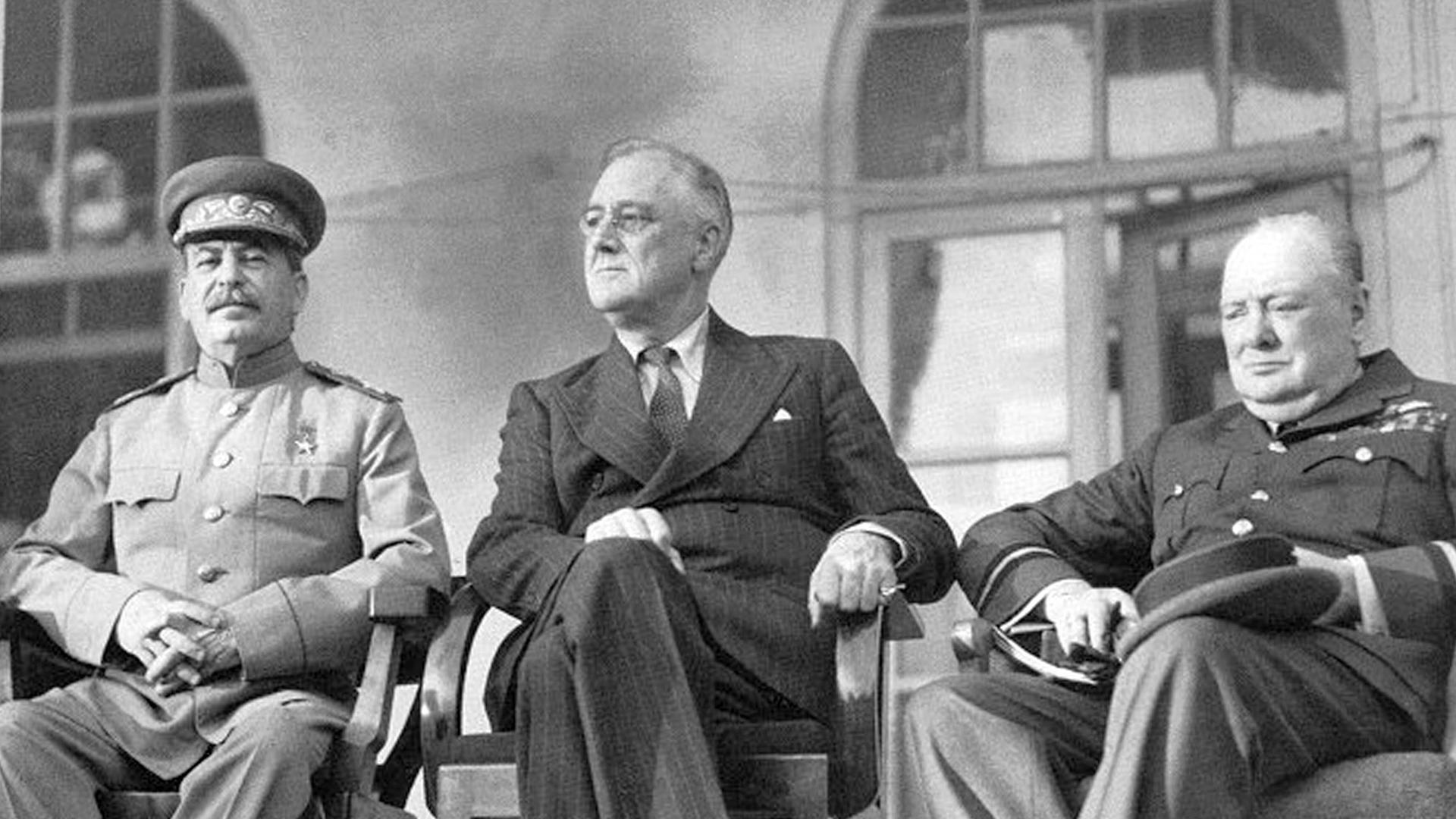 Die Schwarzweißfotografie zeigt Josef Stalin, Franklin D. Roosevelt und Winston Churchill auf Stühlen sitzend auf einer Empore. 