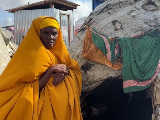 Eine Frau steht in einer gelben Hijab vor einem Zelt in einem Camp und schaut ernst in die Kamera