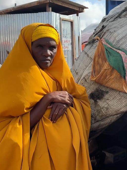 Eine Frau steht in einer gelben Hijab vor einem Zelt in einem Camp und schaut ernst in die Kamera