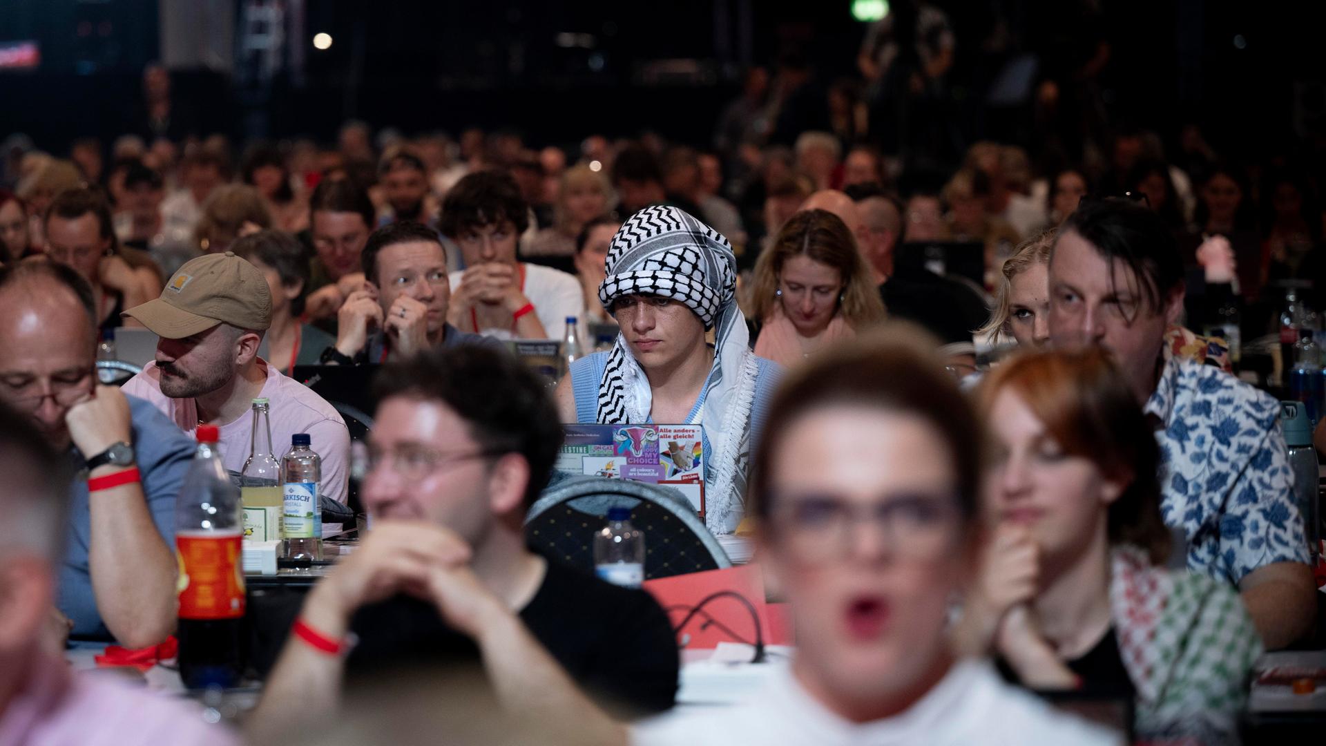 Ein Delegierter auf dem Parteitag der Linken sitzt unter Parteigenossen und hat ein Palästinensertuch als Symbol der Solidarität mit Palästina um seinen Kopf gebunden. Ein Delegierter auf dem Parteitag der Linken sitzt unter Parteigenossen und hat ein Palästinensertuch als Symbol der Solidarität mit Palästina um seinen Kopf gebunden.